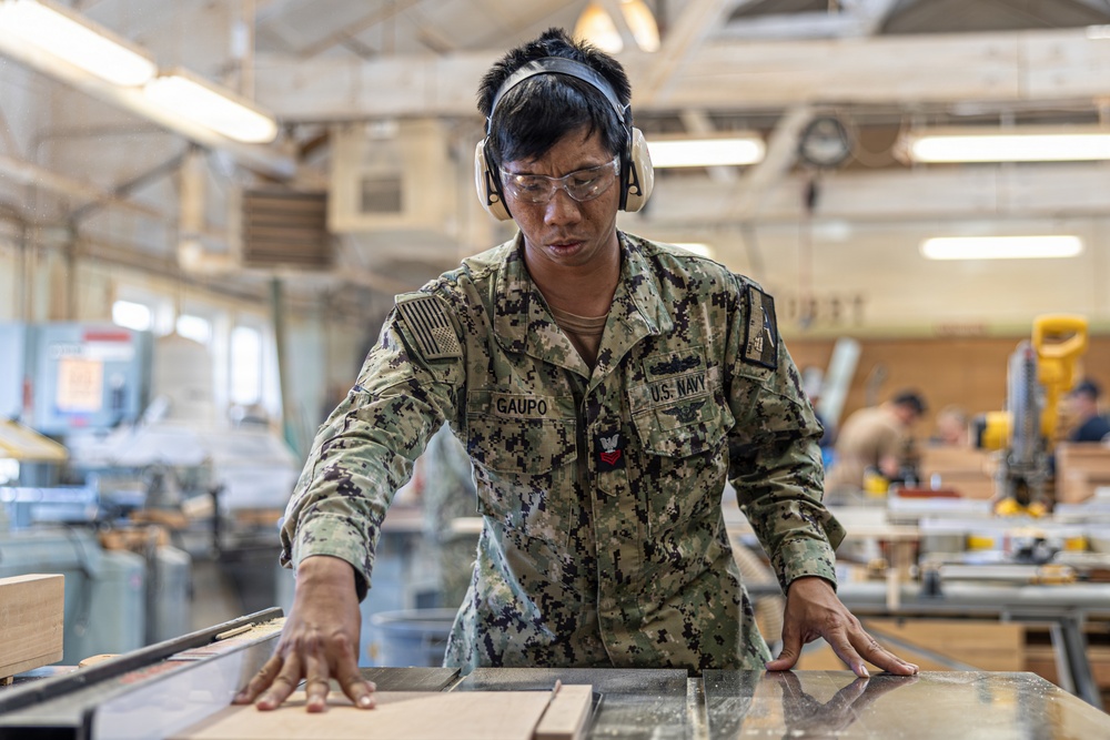 A U.S. Navy service member named Gaupo, wearing NWU (Navy Working Uniform) camouflage, is operating woodworking equipment in what appears to be a military workshop or training facility. The sailor is equipped with proper personal protective equipment including safety glasses and hearing protection earmuffs. Other personnel and woodworking machinery are visible in the background.