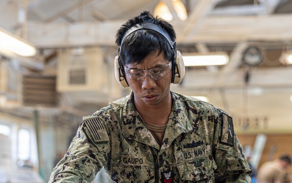 A U.S. Navy service member named Gaupo, wearing NWU (Navy Working Uniform) camouflage, is operating woodworking equipment in what appears to be a military workshop or training facility. The sailor is equipped with proper personal protective equipment including safety glasses and hearing protection earmuffs. Other personnel and woodworking machinery are visible in the background.