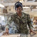 A U.S. Navy service member named Gaupo, wearing NWU (Navy Working Uniform) camouflage, is operating woodworking equipment in what appears to be a military workshop or training facility. The sailor is equipped with proper personal protective equipment including safety glasses and hearing protection earmuffs. Other personnel and woodworking machinery are visible in the background.