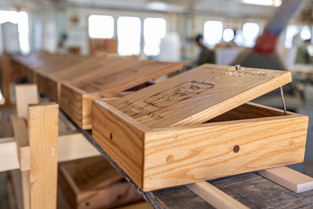 The image shows handcrafted wooden boxes with laser-engraved or carved lids displayed on a workbench in what appears to be a woodworking workshop. The foreground box has its lid propped open, revealing engraved imagery and text on the lid's interior surface. Multiple similar boxes are visible receding into the background of the workshop space.