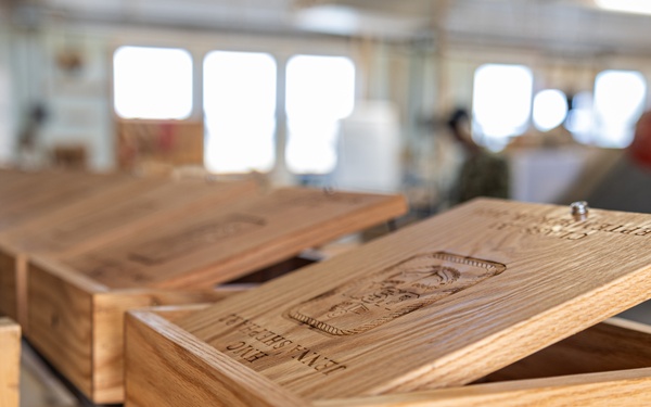 The image shows handcrafted wooden boxes with laser-engraved or carved lids displayed on a workbench in what appears to be a woodworking workshop. The foreground box has its lid propped open, revealing engraved imagery and text on the lid's interior surface. Multiple similar boxes are visible receding into the background of the workshop space.