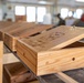 The image shows handcrafted wooden boxes with laser-engraved or carved lids displayed on a workbench in what appears to be a woodworking workshop. The foreground box has its lid propped open, revealing engraved imagery and text on the lid's interior surface. Multiple similar boxes are visible receding into the background of the workshop space.