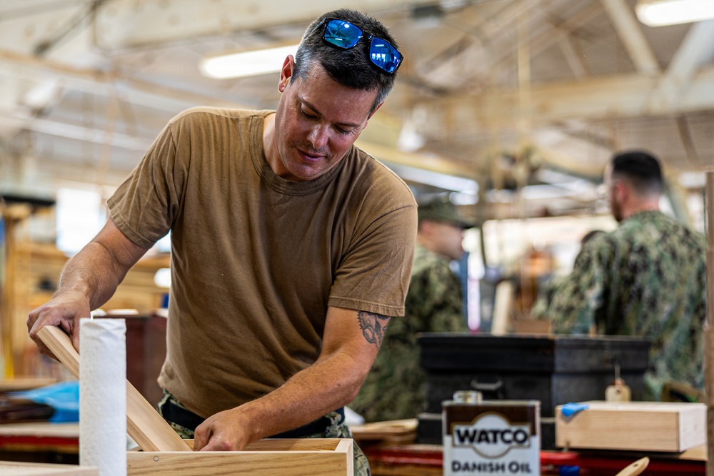 A man in a tan/olive t-shirt with sunglasses on his head works with wooden components in what appears to be a military woodworking shop. Other personnel in camouflage uniforms are visible in the background. A can of Watco Danish Oil wood finish is prominently visible on the workbench.