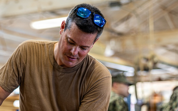 A man in a tan/olive t-shirt with sunglasses on his head works with wooden components in what appears to be a military woodworking shop. Other personnel in camouflage uniforms are visible in the background. A can of Watco Danish Oil wood finish is prominently visible on the workbench.