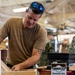 A man in a tan/olive t-shirt with sunglasses on his head works with wooden components in what appears to be a military woodworking shop. Other personnel in camouflage uniforms are visible in the background. A can of Watco Danish Oil wood finish is prominently visible on the workbench.