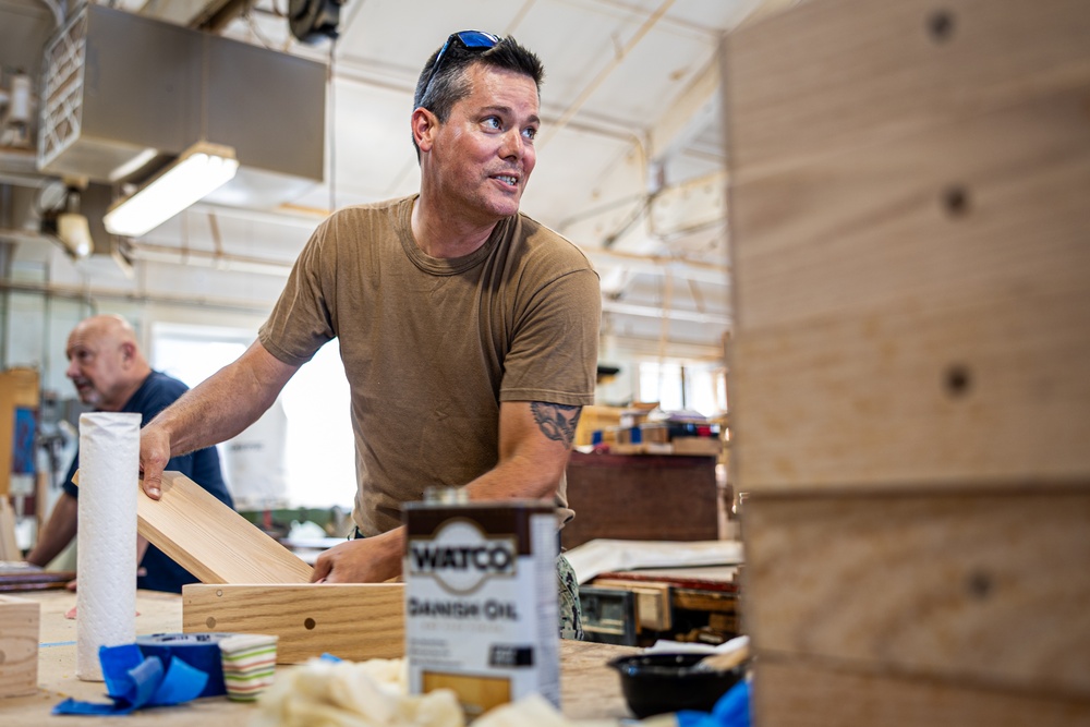 A man in a tan t-shirt with sunglasses on his head works at a woodworking bench in what appears to be a workshop or makerspace. He is handling wooden pieces while a can of Watco Danish Oil and paper towels are visible on the workbench. A second person is partially visible in the background, also engaged in workshop activities.