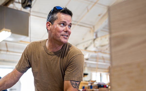 A man in a tan t-shirt with sunglasses on his head works at a woodworking bench in what appears to be a workshop or makerspace. He is handling wooden pieces while a can of Watco Danish Oil and paper towels are visible on the workbench. A second person is partially visible in the background, also engaged in workshop activities.