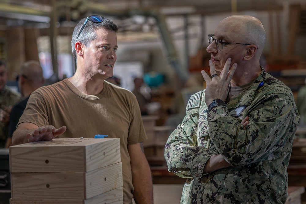 Two men are engaged in a conversation in what appears to be a military workshop or fabrication facility. One man is wearing a tan t-shirt with sunglasses on his head, while the other is in U.S. Navy digital camouflage (NWU Type III) uniform with glasses. They appear to be discussing wooden boxes or crates stacked in the foreground.