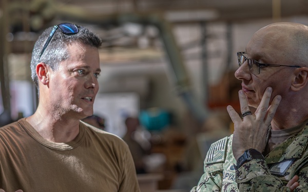 Two men are engaged in a conversation in what appears to be a military workshop or fabrication facility. One man is wearing a tan t-shirt with sunglasses on his head, while the other is in U.S. Navy digital camouflage (NWU Type III) uniform with glasses. They appear to be discussing wooden boxes or crates stacked in the foreground.