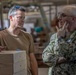 Two men are engaged in a conversation in what appears to be a military workshop or fabrication facility. One man is wearing a tan t-shirt with sunglasses on his head, while the other is in U.S. Navy digital camouflage (NWU Type III) uniform with glasses. They appear to be discussing wooden boxes or crates stacked in the foreground.