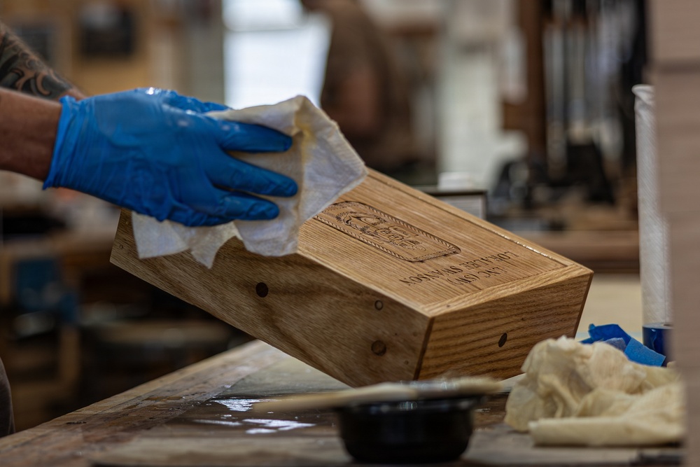 A craftsperson wearing blue nitrile gloves is wiping or finishing a wooden box with a cloth or paper towel in what appears to be a woodworking workshop. The wooden box has a laser-engraved logo and text on its surface. The workbench shows signs of active use with finishing supplies visible nearby.
