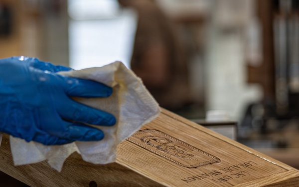 A craftsperson wearing blue nitrile gloves is wiping or finishing a wooden box with a cloth or paper towel in what appears to be a woodworking workshop. The wooden box has a laser-engraved logo and text on its surface. The workbench shows signs of active use with finishing supplies visible nearby.