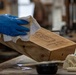 A craftsperson wearing blue nitrile gloves is wiping or finishing a wooden box with a cloth or paper towel in what appears to be a woodworking workshop. The wooden box has a laser-engraved logo and text on its surface. The workbench shows signs of active use with finishing supplies visible nearby.