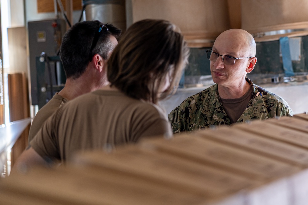 Three individuals are engaged in a conversation in what appears to be an indoor workspace or facility. One person on the right is wearing U.S. Navy digital camouflage (NWU Type III) uniform with glasses and a shaved head. The other two individuals, viewed from behind, are wearing tan/khaki civilian or military-style shirts.