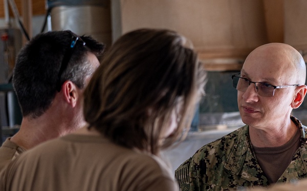 Three individuals are engaged in a conversation in what appears to be an indoor workspace or facility. One person on the right is wearing U.S. Navy digital camouflage (NWU Type III) uniform with glasses and a shaved head. The other two individuals, viewed from behind, are wearing tan/khaki civilian or military-style shirts.