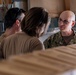Three individuals are engaged in a conversation in what appears to be an indoor workspace or facility. One person on the right is wearing U.S. Navy digital camouflage (NWU Type III) uniform with glasses and a shaved head. The other two individuals, viewed from behind, are wearing tan/khaki civilian or military-style shirts.