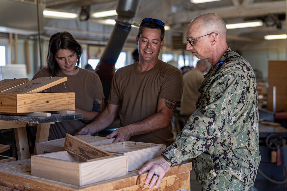 Three individuals are gathered in a woodworking shop examining handcrafted wooden boxes. Two people wearing tan/brown t-shirts and one person in U.S. Navy digital camouflage (NWU Type III) uniform are inspecting the finished wooden pieces. The setting appears to be a military woodworking facility or hobby shop with various woodworking equipment visible in the background.