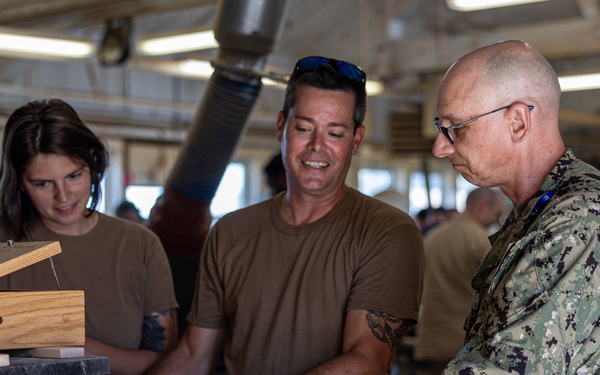 Three individuals are gathered in a woodworking shop examining handcrafted wooden boxes. Two people wearing tan/brown t-shirts and one person in U.S. Navy digital camouflage (NWU Type III) uniform are inspecting the finished wooden pieces. The setting appears to be a military woodworking facility or hobby shop with various woodworking equipment visible in the background.