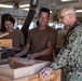 Three individuals are gathered in a woodworking shop examining handcrafted wooden boxes. Two people wearing tan/brown t-shirts and one person in U.S. Navy digital camouflage (NWU Type III) uniform are inspecting the finished wooden pieces. The setting appears to be a military woodworking facility or hobby shop with various woodworking equipment visible in the background.