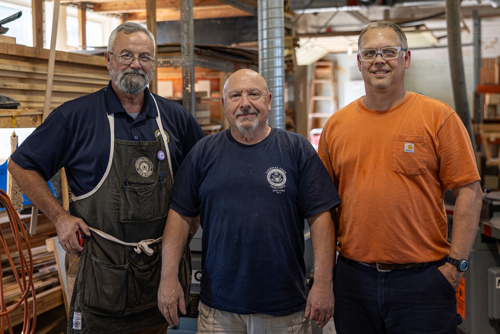 Three middle-aged men pose together for a photo inside what appears to be a woodworking shop or carpentry workspace. The men are dressed in work attire, including an apron, work t-shirts, and safety glasses, suggesting they are tradespeople or craftsmen. The background shows lumber, tools, and workshop equipment consistent with a professional woodworking environment.