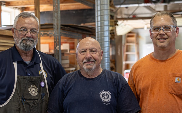 Three middle-aged men pose together for a photo inside what appears to be a woodworking shop or carpentry workspace. The men are dressed in work attire, including an apron, work t-shirts, and safety glasses, suggesting they are tradespeople or craftsmen. The background shows lumber, tools, and workshop equipment consistent with a professional woodworking environment.