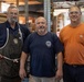 Three middle-aged men pose together for a photo inside what appears to be a woodworking shop or carpentry workspace. The men are dressed in work attire, including an apron, work t-shirts, and safety glasses, suggesting they are tradespeople or craftsmen. The background shows lumber, tools, and workshop equipment consistent with a professional woodworking environment.