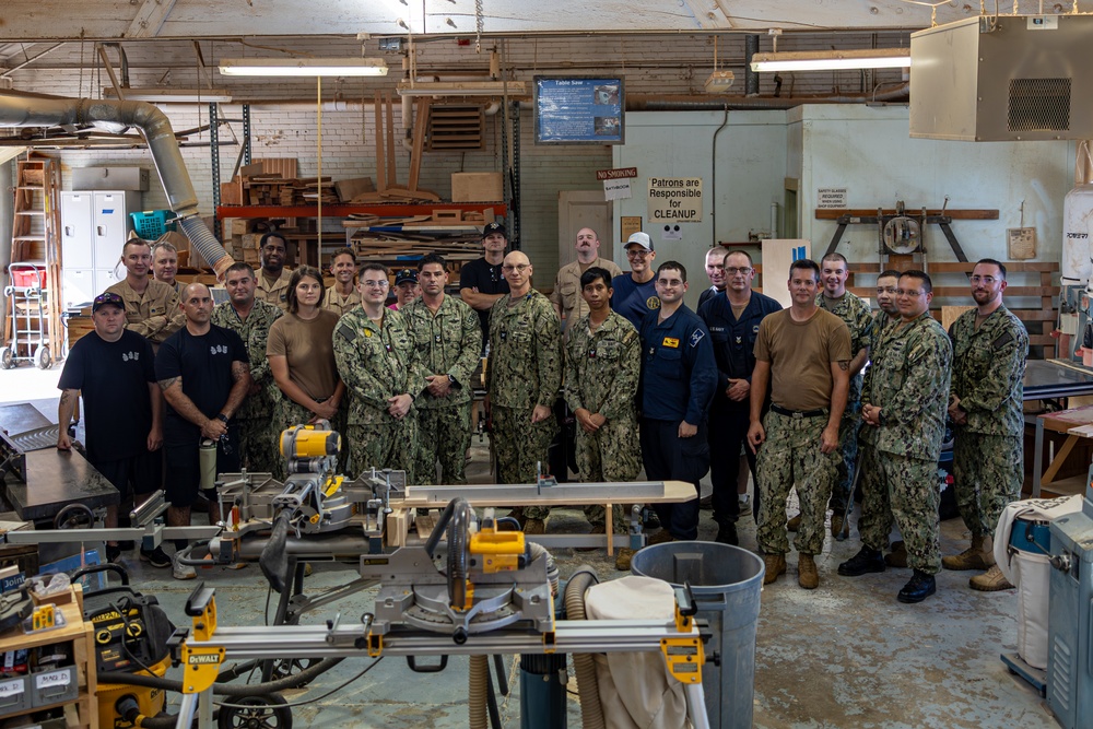 A large group of approximately 25 military personnel, predominantly wearing U.S. Navy NWU (Naval Working Uniform) camouflage pattern uniforms, pose for a group photo inside a woodworking workshop. The facility appears to be a military MWR (Morale, Welfare, and Recreation) or hobby shop equipped with various woodworking tools and machinery. Several civilians in dark clothing are also present among the uniformed personnel.