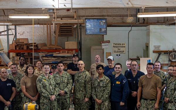 A large group of approximately 25 military personnel, predominantly wearing U.S. Navy NWU (Naval Working Uniform) camouflage pattern uniforms, pose for a group photo inside a woodworking workshop. The facility appears to be a military MWR (Morale, Welfare, and Recreation) or hobby shop equipped with various woodworking tools and machinery. Several civilians in dark clothing are also present among the uniformed personnel.