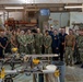 A large group of approximately 25 military personnel, predominantly wearing U.S. Navy NWU (Naval Working Uniform) camouflage pattern uniforms, pose for a group photo inside a woodworking workshop. The facility appears to be a military MWR (Morale, Welfare, and Recreation) or hobby shop equipped with various woodworking tools and machinery. Several civilians in dark clothing are also present among the uniformed personnel.