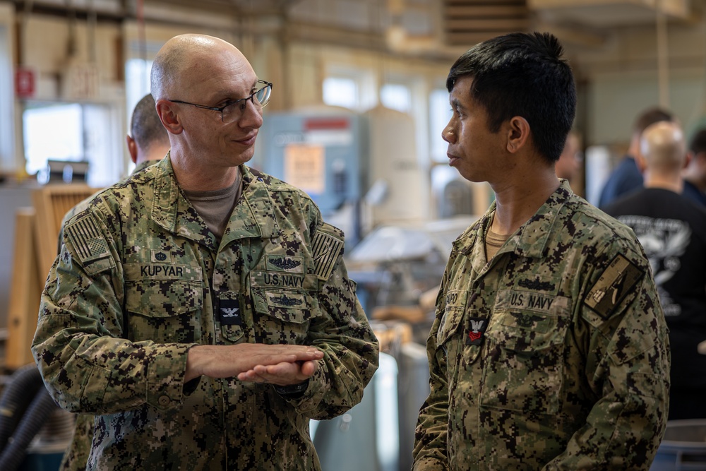 Two U.S. Navy personnel in NWU Type III (woodland digital camouflage) uniforms are engaged in conversation inside what appears to be a workshop or industrial facility. The individual on the left, identified by name tape as 'KUPYAR,' appears to be of higher rank and is gesturing while speaking. A third person is visible in the blurred background.