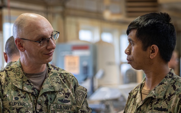 Two U.S. Navy personnel in NWU Type III (woodland digital camouflage) uniforms are engaged in conversation inside what appears to be a workshop or industrial facility. The individual on the left, identified by name tape as 'KUPYAR,' appears to be of higher rank and is gesturing while speaking. A third person is visible in the blurred background.