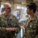 Two U.S. Navy personnel in NWU Type III (woodland digital camouflage) uniforms are engaged in conversation inside what appears to be a workshop or industrial facility. The individual on the left, identified by name tape as 'KUPYAR,' appears to be of higher rank and is gesturing while speaking. A third person is visible in the blurred background.