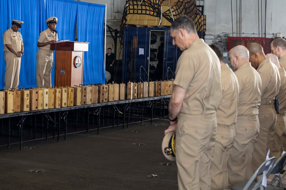 A military ceremony is taking place in what appears to be a ship's hangar bay or large indoor facility. An officer is speaking at a podium adorned with a military insignia, while a row of personnel in tan/khaki uniforms stand at attention. A long table displays numerous identical wooden presentation boxes, suggesting a retirement or award ceremony.