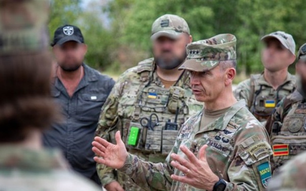 A group of military personnel in multicam/OCP uniforms are gathered in an outdoor briefing or discussion. A central figure wearing a four-star rank insignia on his cap is speaking and gesturing with both hands to the assembled group. Several individuals in the background have their faces blurred, and Ukrainian flag patches are visible on some uniforms.