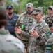 A group of military personnel in multicam/OCP uniforms are gathered in an outdoor briefing or discussion. A central figure wearing a four-star rank insignia on his cap is speaking and gesturing with both hands to the assembled group. Several individuals in the background have their faces blurred, and Ukrainian flag patches are visible on some uniforms.