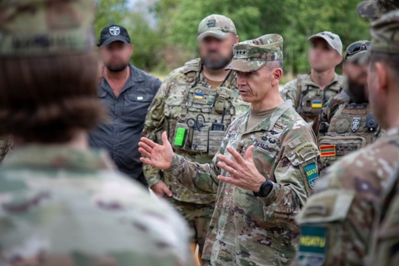 A group of military personnel in multicam/OCP uniforms are gathered in an outdoor briefing or discussion. A central figure wearing a four-star rank insignia on his cap is speaking and gesturing with both hands to the assembled group. Several individuals in the background have their faces blurred, and Ukrainian flag patches are visible on some uniforms.