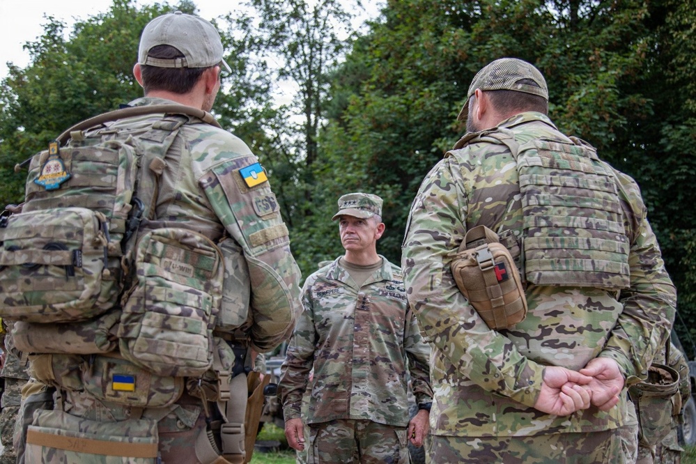 Three military personnel in camouflage uniforms are engaged in a conversation outdoors near a tree line. The central figure wears a standard military patrol cap and appears to be a senior officer based on rank insignia visible on the collar area. Two individuals facing away from the camera wear tactical gear with Ukrainian flag patches visible on their sleeves.