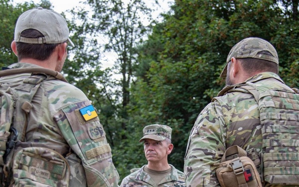 Three military personnel in camouflage uniforms are engaged in a conversation outdoors near a tree line. The central figure wears a standard military patrol cap and appears to be a senior officer based on rank insignia visible on the collar area. Two individuals facing away from the camera wear tactical gear with Ukrainian flag patches visible on their sleeves.