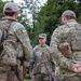 Three military personnel in camouflage uniforms are engaged in a conversation outdoors near a tree line. The central figure wears a standard military patrol cap and appears to be a senior officer based on rank insignia visible on the collar area. Two individuals facing away from the camera wear tactical gear with Ukrainian flag patches visible on their sleeves.
