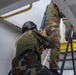 The image shows multiple armed personnel in camouflage uniforms ascending a metal staircase or ladder on what appears to be a vessel or industrial structure. The individuals are equipped with body armor, helmets, and rifles, suggesting a tactical boarding or clearance operation. The scene appears to be a training exercise or operational boarding scenario.