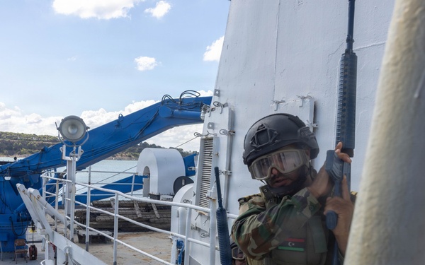 The image shows armed military personnel in camouflage uniforms conducting what appears to be a maritime boarding or vessel clearance exercise on the deck of a ship. The lead soldier is wearing a tactical helmet with goggles and carrying a rifle, with a flag patch visible on his shoulder. Additional armed personnel are visible in the background on the ship's deck.