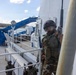 The image shows armed military personnel in camouflage uniforms conducting what appears to be a maritime boarding or vessel clearance exercise on the deck of a ship. The lead soldier is wearing a tactical helmet with goggles and carrying a rifle, with a flag patch visible on his shoulder. Additional armed personnel are visible in the background on the ship's deck.