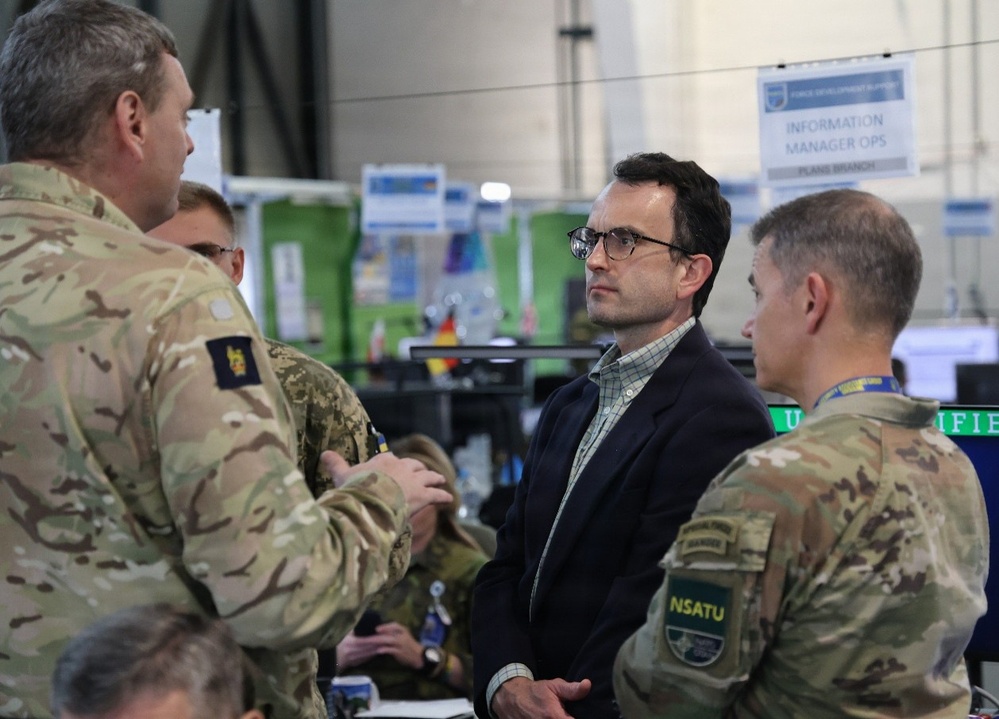 The image shows a briefing or discussion taking place in what appears to be a military operations center or command post. Several military personnel in camouflage uniforms are engaged in conversation with a civilian man wearing glasses and a dark blazer. Signage in the background indicates this is related to an 'Information Manager Ops / Plans Branch' function.