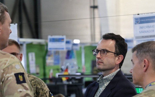 The image shows a briefing or discussion taking place in what appears to be a military operations center or command post. Several military personnel in camouflage uniforms are engaged in conversation with a civilian man wearing glasses and a dark blazer. Signage in the background indicates this is related to an 'Information Manager Ops / Plans Branch' function.