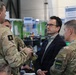 The image shows a briefing or discussion taking place in what appears to be a military operations center or command post. Several military personnel in camouflage uniforms are engaged in conversation with a civilian man wearing glasses and a dark blazer. Signage in the background indicates this is related to an 'Information Manager Ops / Plans Branch' function.