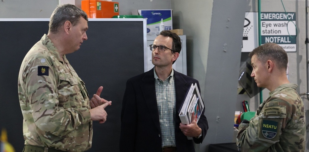 Three individuals are engaged in a discussion inside what appears to be a military facility or workshop. Two are wearing military camouflage uniforms while one civilian in a blazer holds binders or folders. The setting includes safety signage and storage equipment typical of a military technical or maintenance environment.