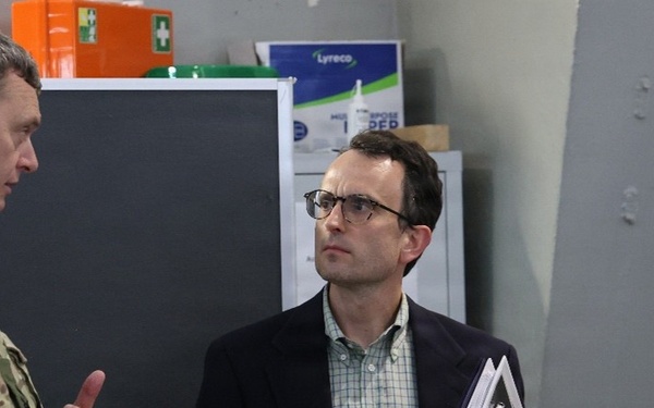 Three individuals are engaged in a discussion inside what appears to be a military facility or workshop. Two are wearing military camouflage uniforms while one civilian in a blazer holds binders or folders. The setting includes safety signage and storage equipment typical of a military technical or maintenance environment.