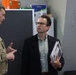 Three individuals are engaged in a discussion inside what appears to be a military facility or workshop. Two are wearing military camouflage uniforms while one civilian in a blazer holds binders or folders. The setting includes safety signage and storage equipment typical of a military technical or maintenance environment.