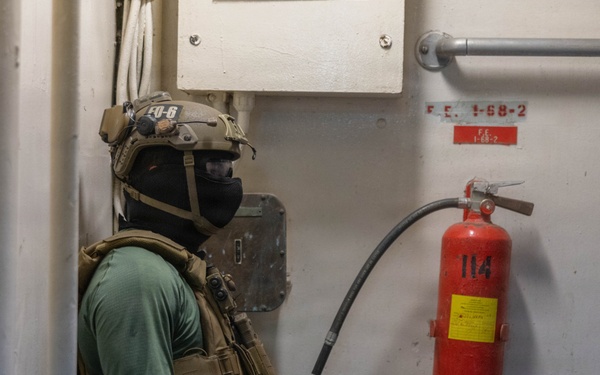 A heavily equipped tactical operator in full combat gear is seated on the floor of what appears to be a naval vessel corridor or compartment. The individual is wearing a tactical helmet, balaclava, plate carrier vest, knee pads, and combat boots. A red fire extinguisher labeled '114' is mounted on the wall nearby, with a placard reading 'F.E. 1-68-2'.