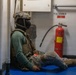 A heavily equipped tactical operator in full combat gear is seated on the floor of what appears to be a naval vessel corridor or compartment. The individual is wearing a tactical helmet, balaclava, plate carrier vest, knee pads, and combat boots. A red fire extinguisher labeled '114' is mounted on the wall nearby, with a placard reading 'F.E. 1-68-2'.